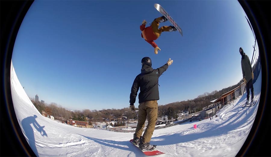 Snowboarder goes off jump and gives his friend a High 5 in mid-air.