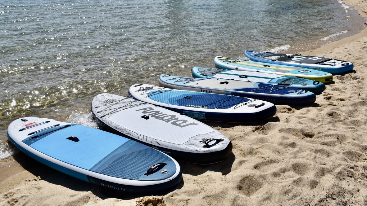 a lineup of seven inflatable paddle boards being tested side-by-side on the beach at lake tahoe