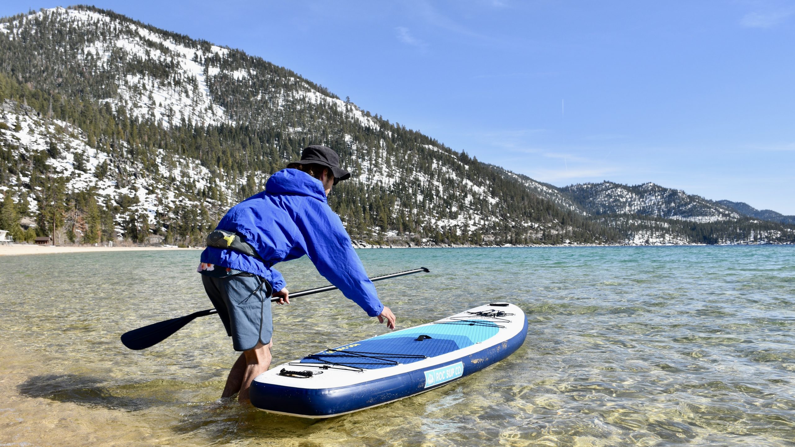 a man launching the roc kahuna inflatable paddle board into lake tahoe with mountains in the background