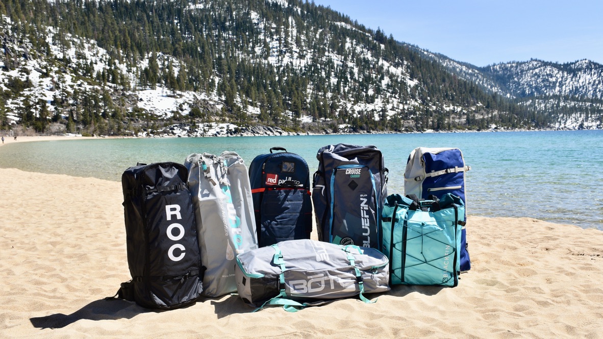 a lineup of seven inflatable paddle board duffel bags on the beach at lake tahoe with mountains in the background