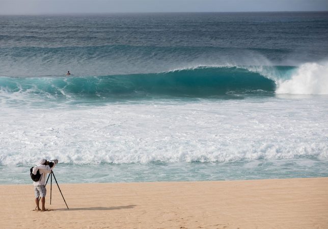 a photographer on the beach in maui shooting towards an empty barreling wave with a surfer in the water