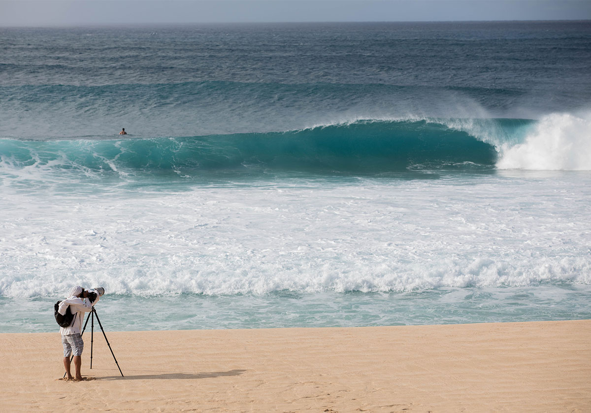 a photographer on the beach in maui shooting towards an empty barreling wave with a surfer in the water