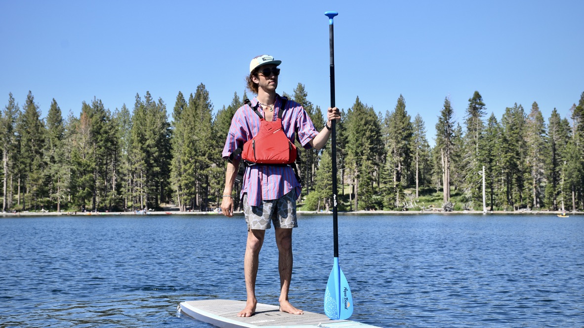 A man standing on a paddle board on an alpine lake with trees in the background
