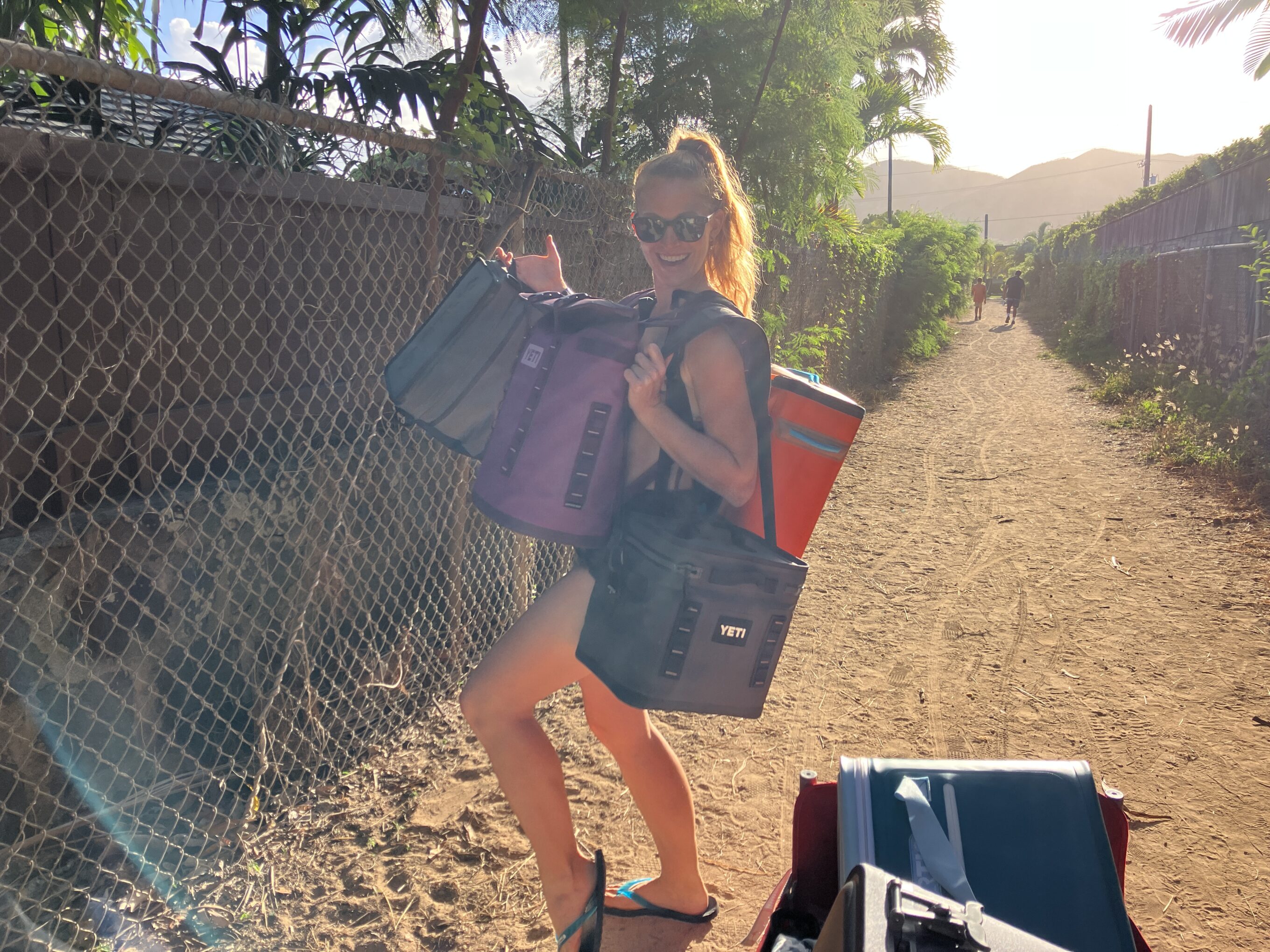 woman walking down a dirt path carrying a bunch of soft coolers