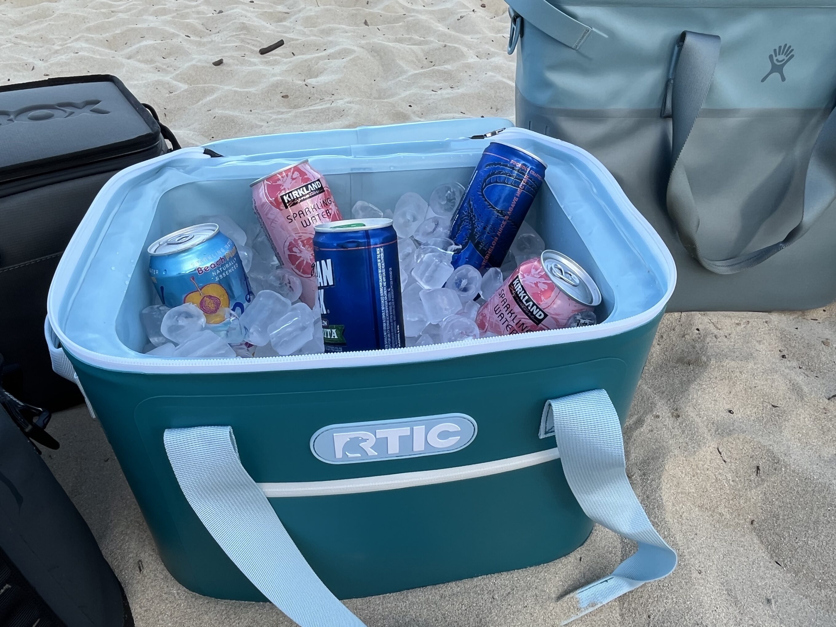 an RTIC soft cooler sitting open on the beach with drinks and a bunch of ice in it
