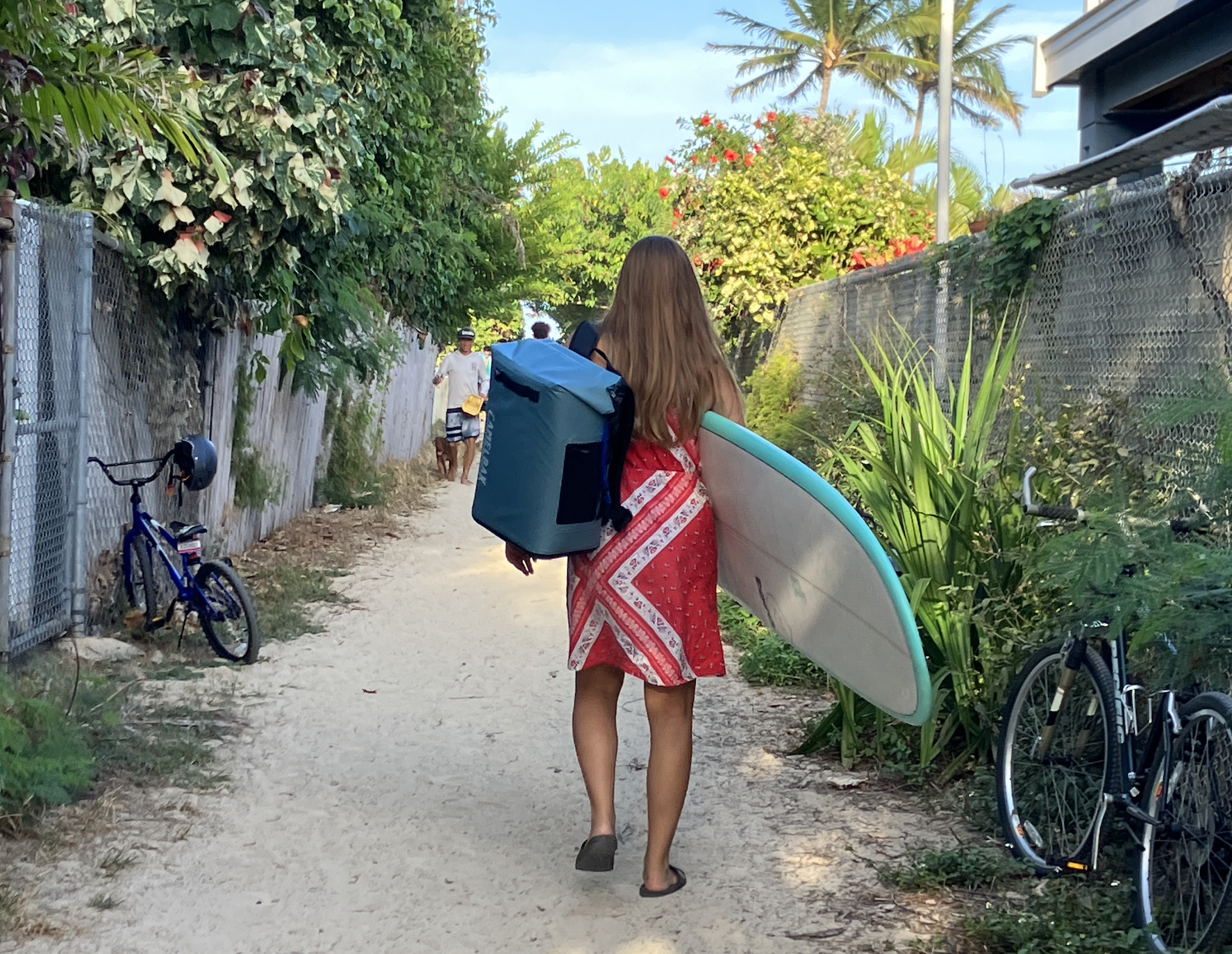 women walking down a dirt path with a surfboard in one arm and a backpack cooler in the other