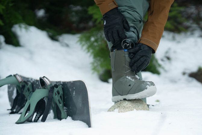 A snowboarder putting on boots next to his snowboard