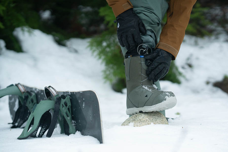 A snowboarder putting on boots next to his snowboard