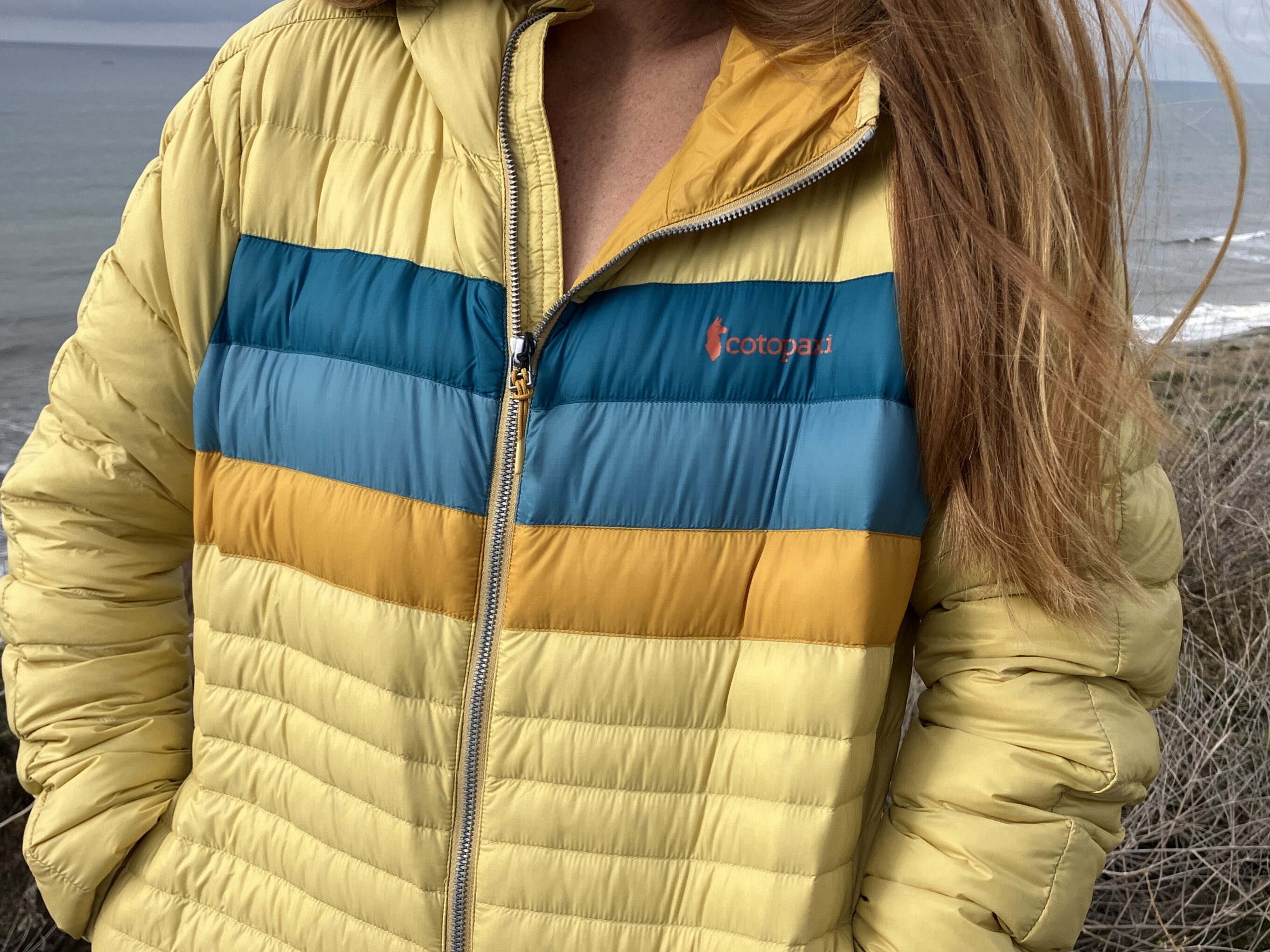 A woman wears the Cotopaxi Fuego Down Hoodie with the beach in the background
