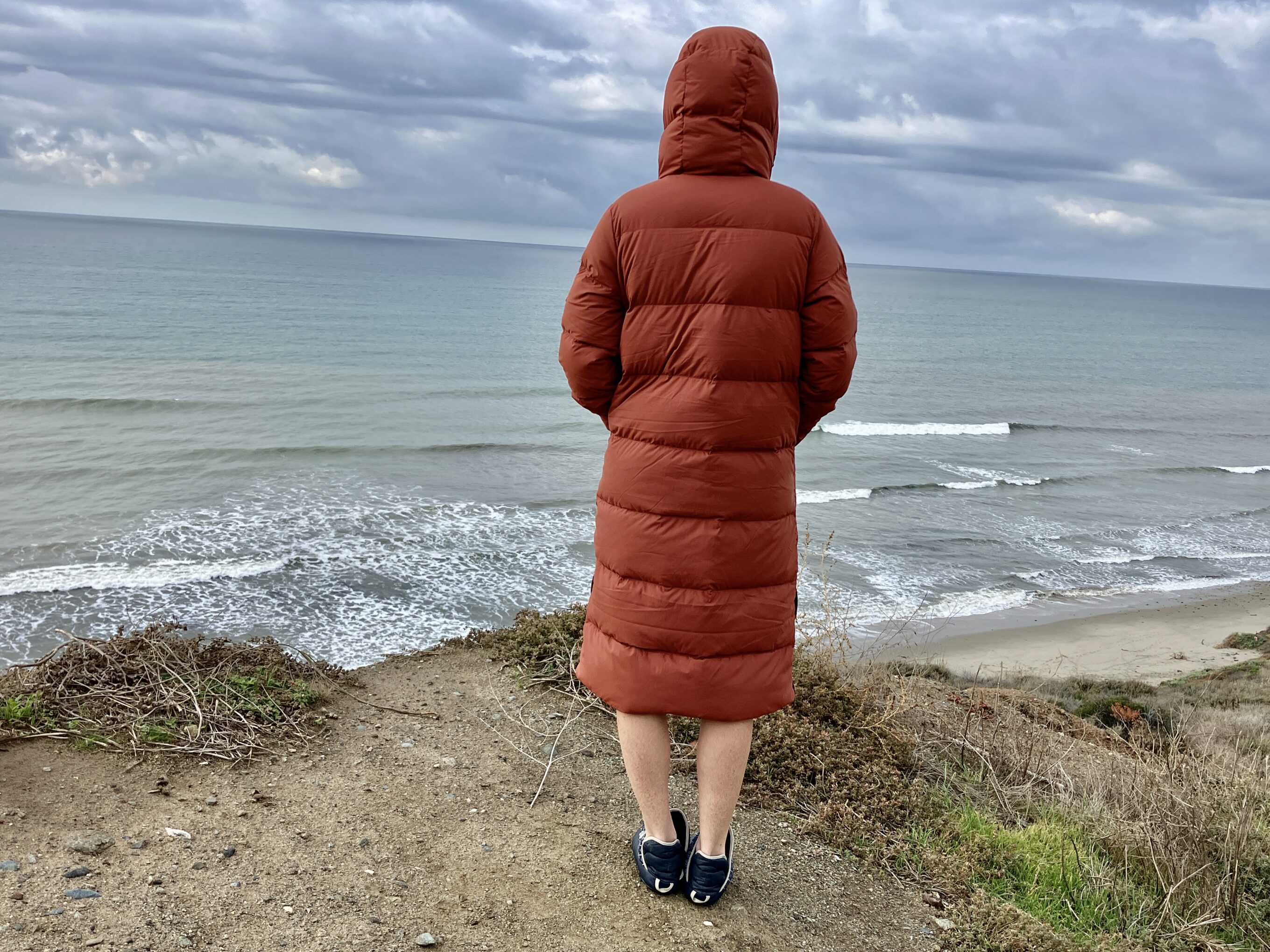 a women facing the ocean at the beach while wearing the Outdoor Research Coze Down Parka