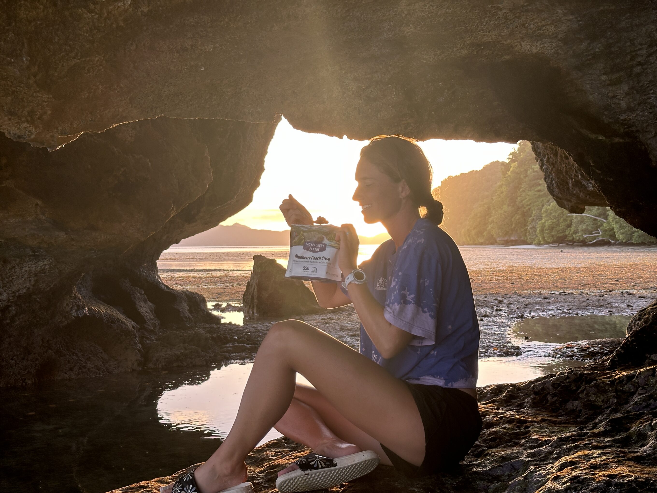 A woman eats one of the backpacking meals from this guide while sitting in a cave at the beach