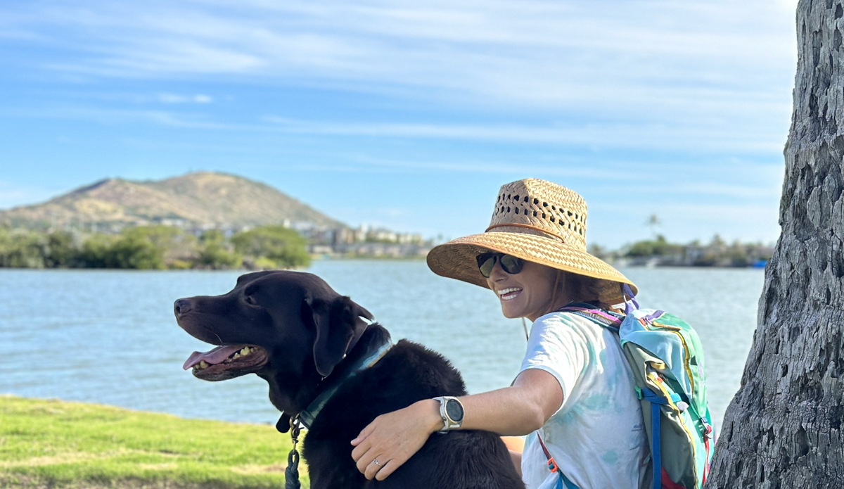 woman in a sun hat sitting by the water with a dog