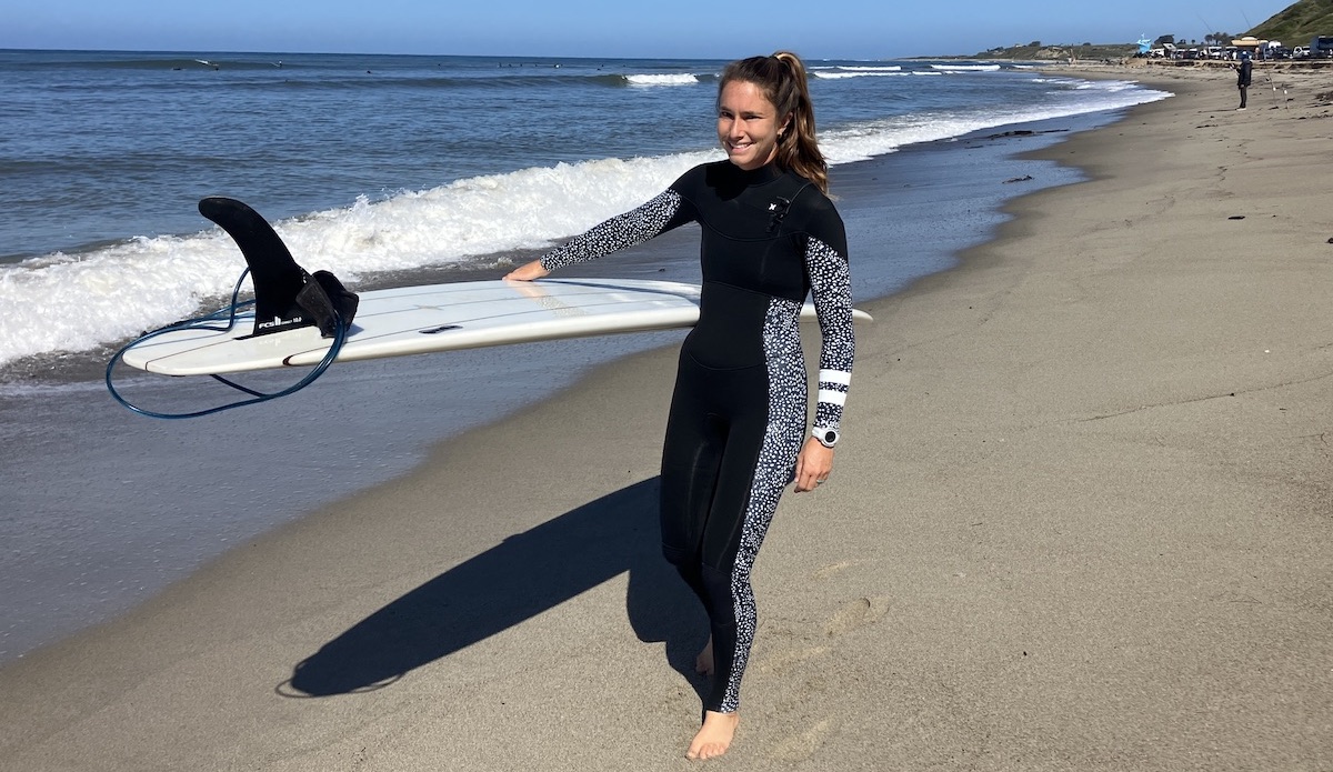 a woman wearing a hurley wetsuit walking on the beach with a surfboard