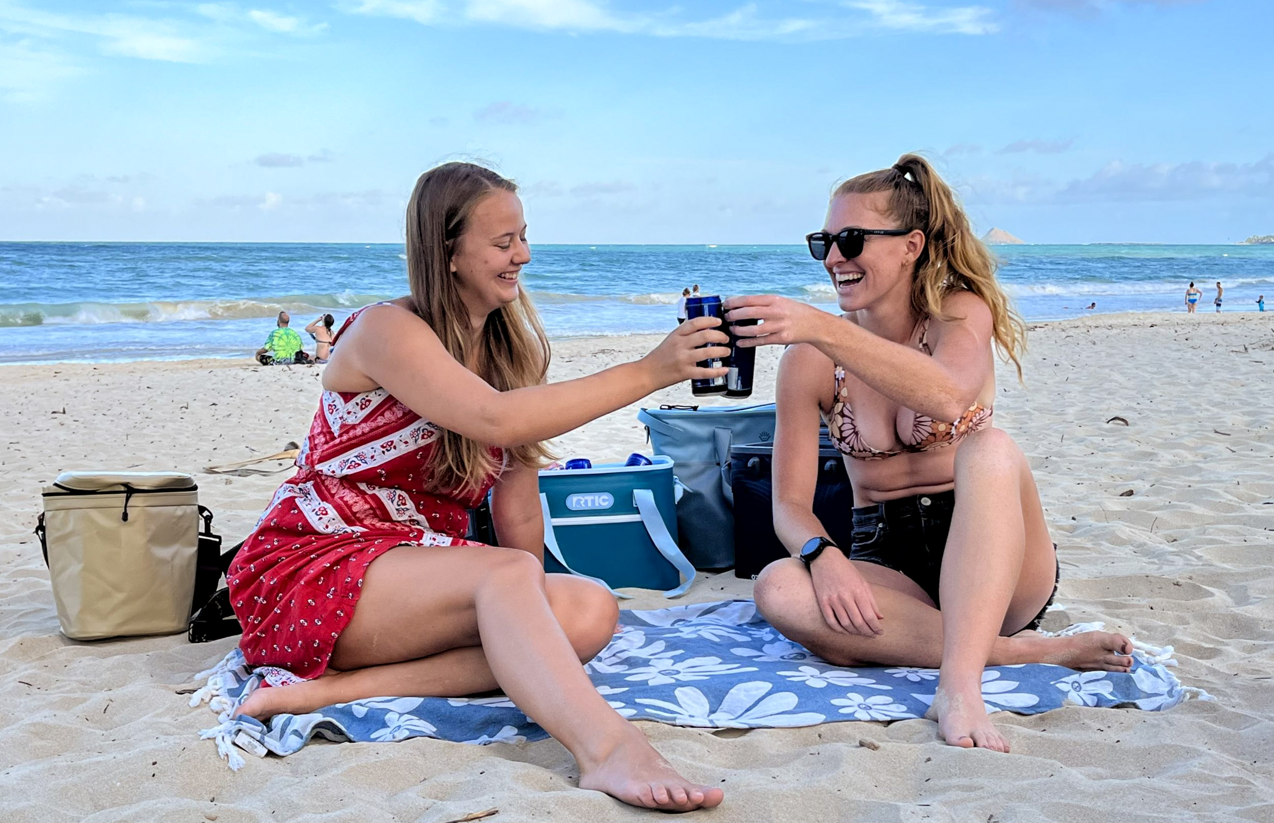 two women enjoying a drink on the beach in front of soft coolers