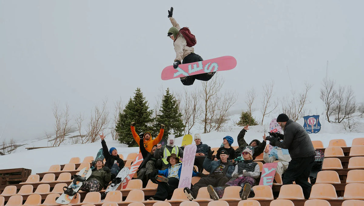 a group of snowboarders sit under a snowborder jumping over them doing a method grab