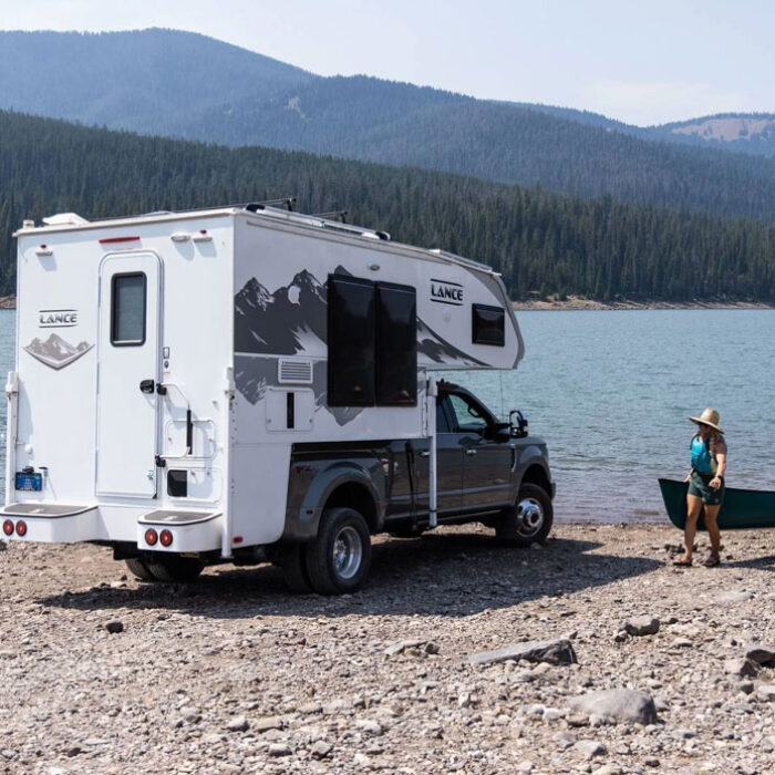 A view of a truck camper and people carrying a canoe towards a lake in the mountains