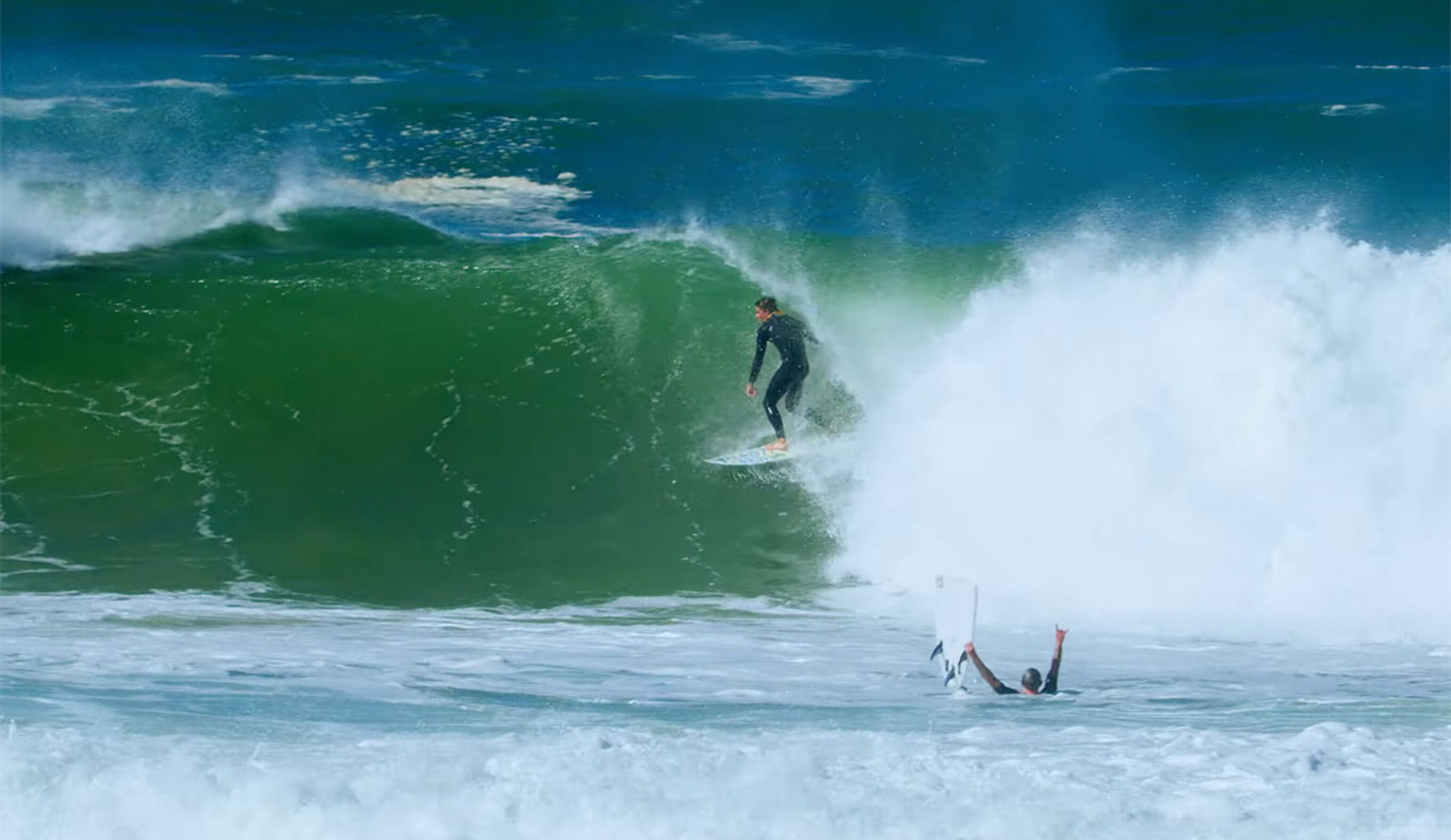 Surfer exiting barrel while another surfer celebrates with a broken surfboard