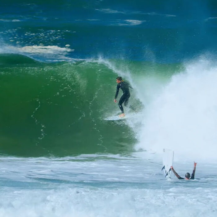 Surfer exiting barrel while another surfer celebrates with a broken surfboard