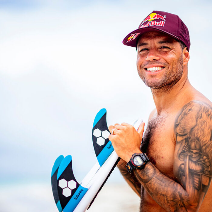 Adriano de Souza smiling with a Red Bull hat while leaning on a surfboard