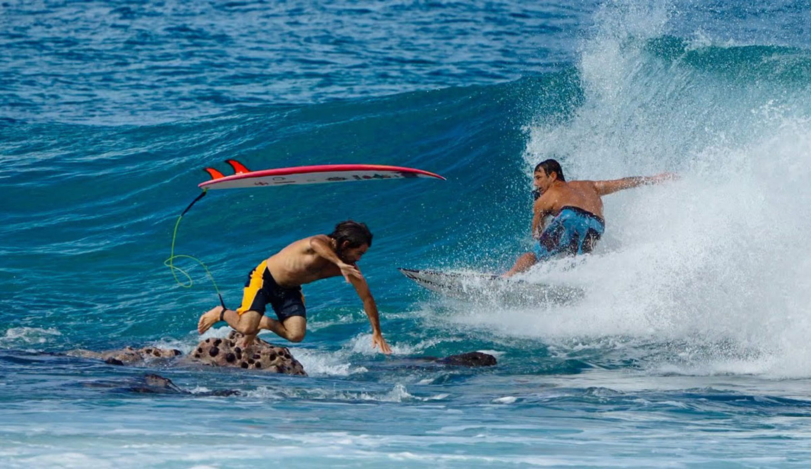 Mason Ho jumping off a rock as Sheldon Paishon surfs past him